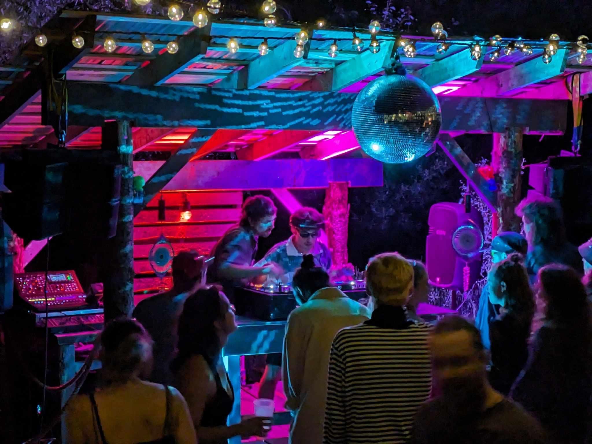 Vue sur un DJ booth avec 2 DJ à la console, éléments d'éclairage de scène extérieure, énorme boule disco au-dessus et plusieurs danseurs prêts du DJ booth, sur fonds d'éclairage nocturne rose, mauve et bleu
