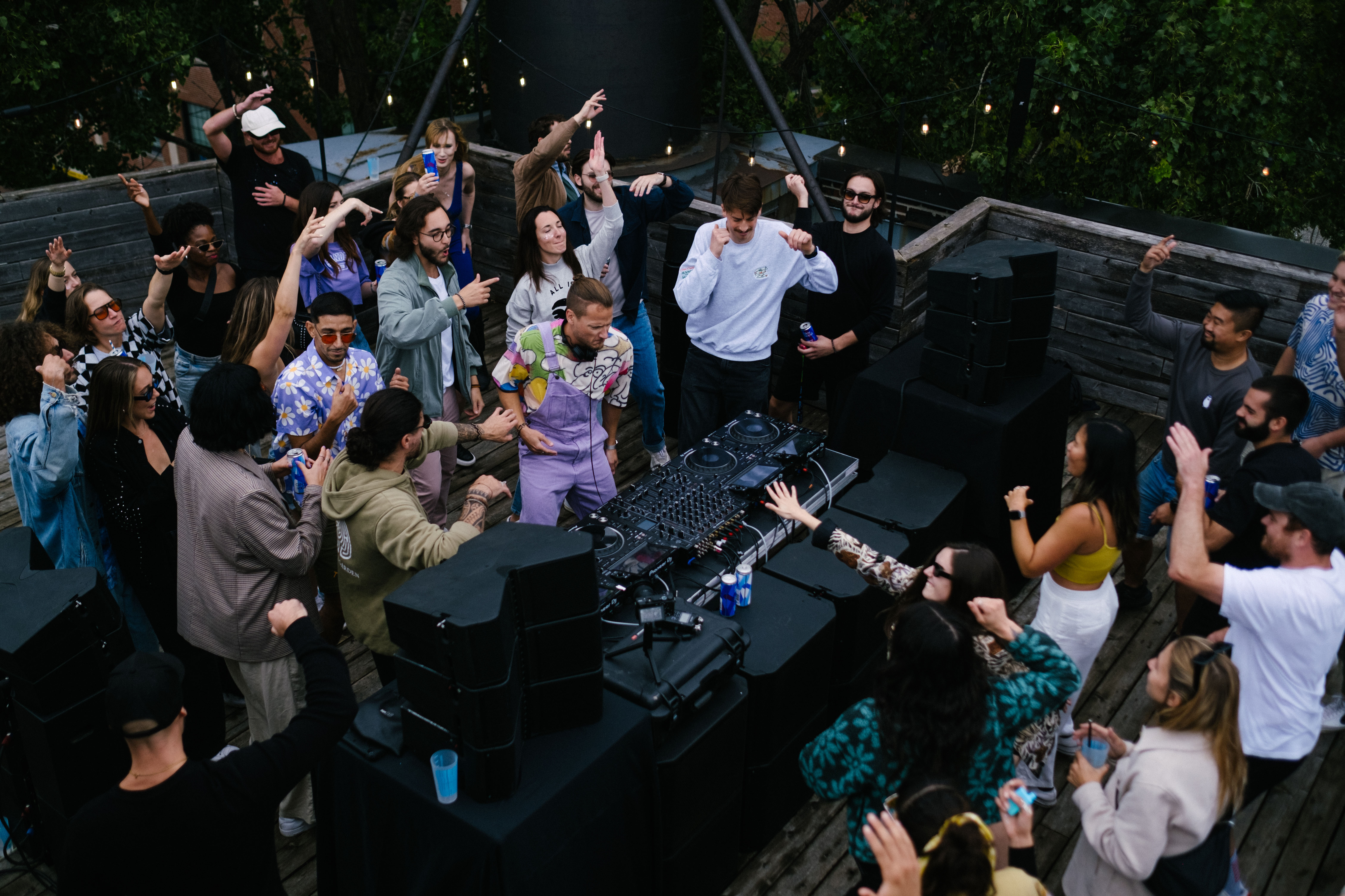 Un party sur une terrasse extérieure, avec un DJ booth et un DJ en pleine performance, entourés de danseurs et un équipement de sonorisation impressionnant avec tables tournantes, haut-parleurs, console...