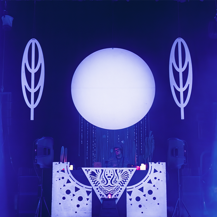 Ambiance bleutée, DJ booth et DJ en pleine prestation, sous un énorme globe lumineux blanc, avec éléments de décors blancs en forme de feuille à gauche et à droite, décoration comportant des triangles dans le DJ booth
