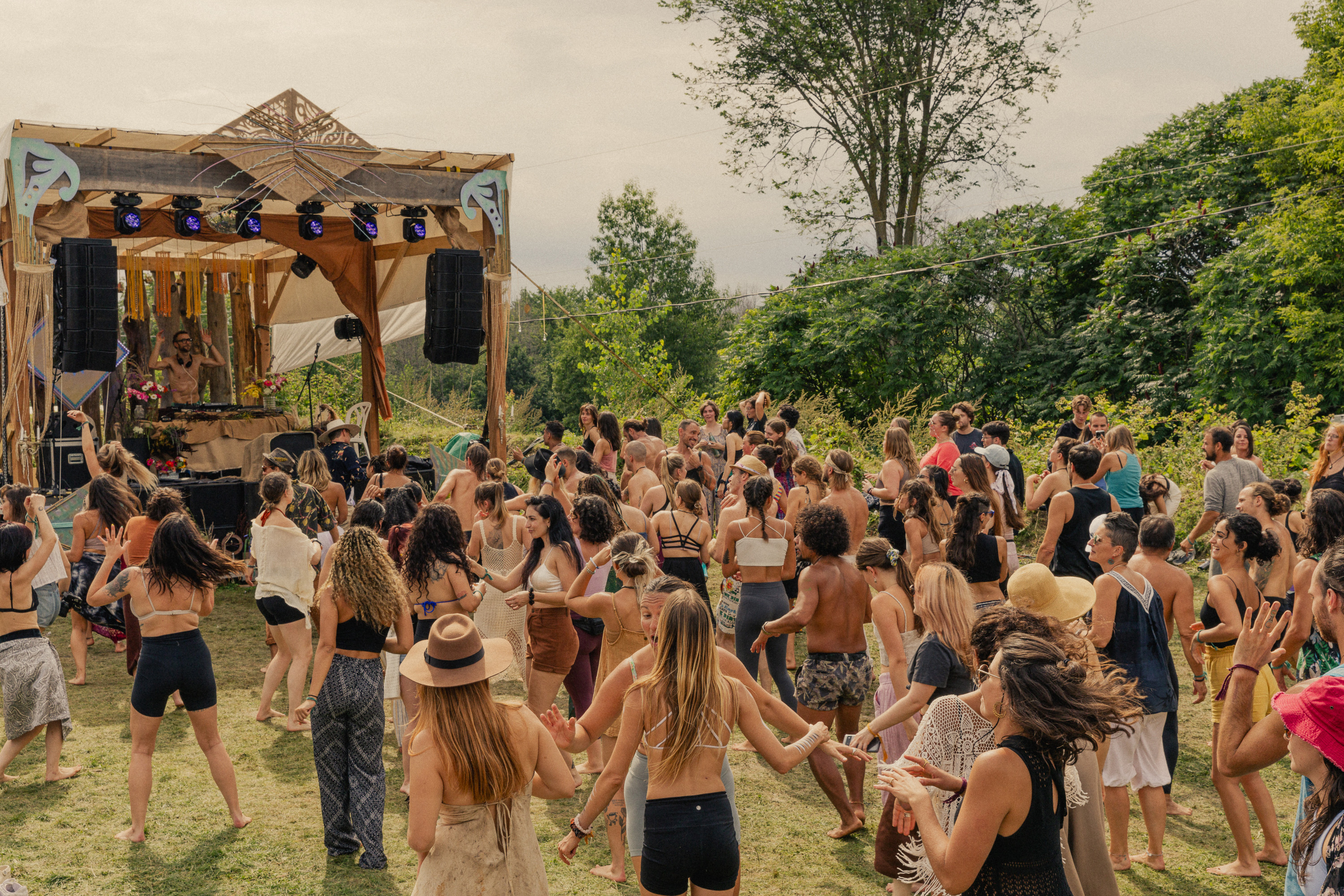 Une foule de jeunes danseurs sur une piste de danse extérieur durant un événement estival devant une scène comportant un DJ booth, des haut-parleurs superposés et l'équipement d'éclairage en pleine nature