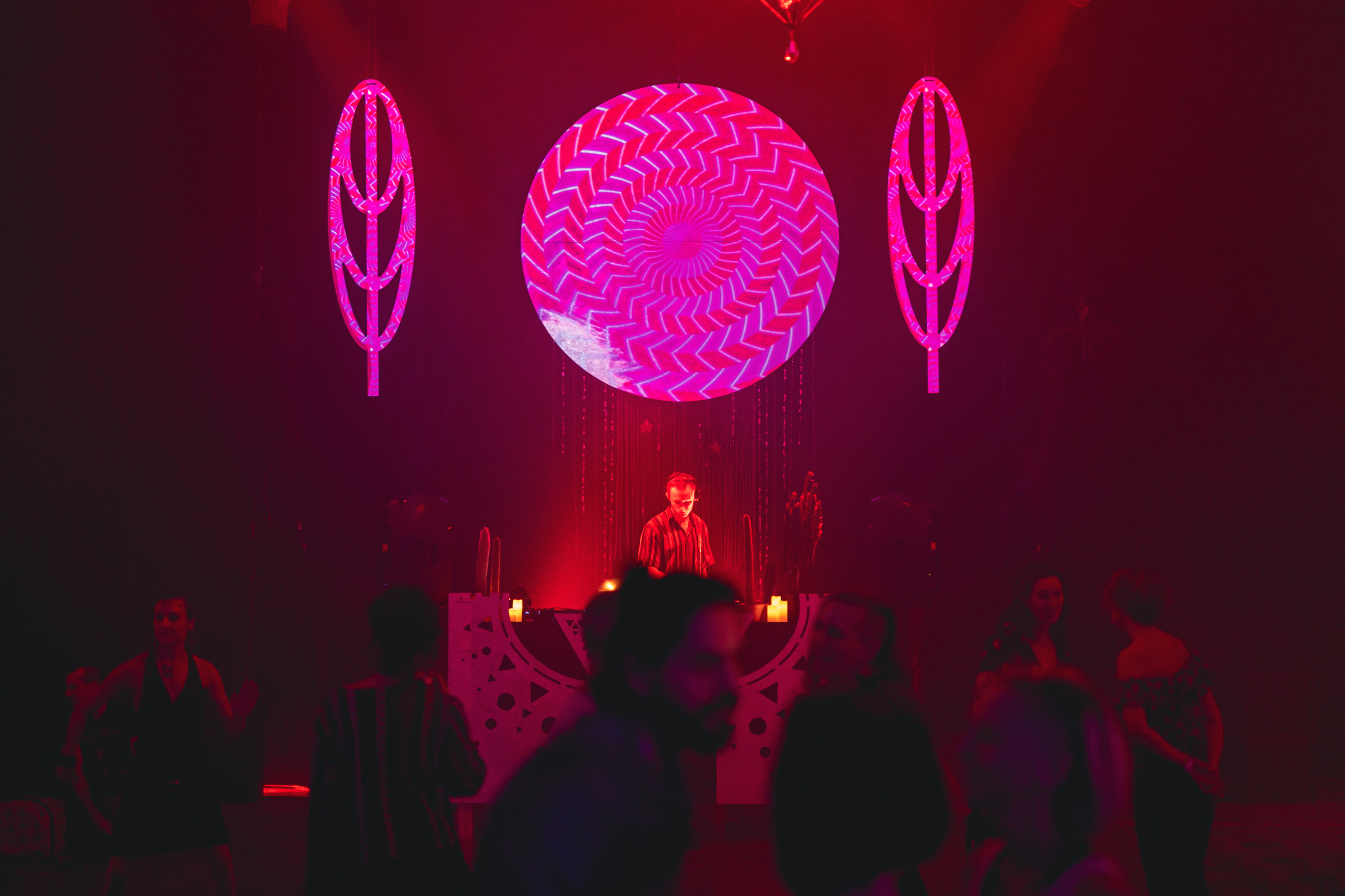 UN DJ en pleine performance durant un événement intérieur, sur fonds de tientes roses et rouge profond, avec une boule géante lumineuse au-dessus et entouré de deux éléments de décor en forme de feuilles teintés rose, avec des danseurs sur la piste de danse