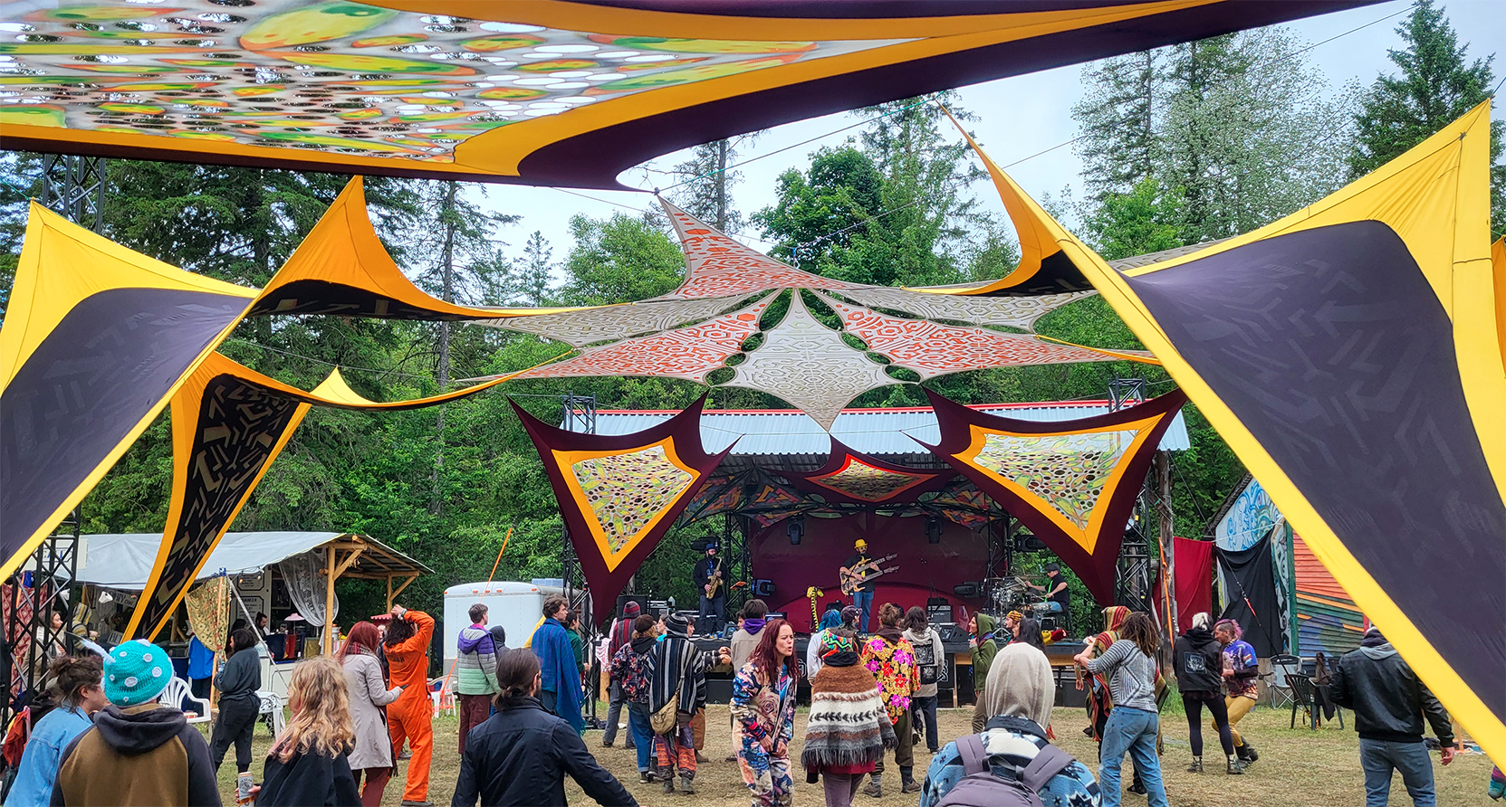 Piste de danse extérieure d'un festival de musique électronique en pleine nature, décoration avec tissus jaunes et mauves, vibrants, tendus au dessus des danseurs qui assistent à une prestation musicale d'un groupe d'une la scène adjacente