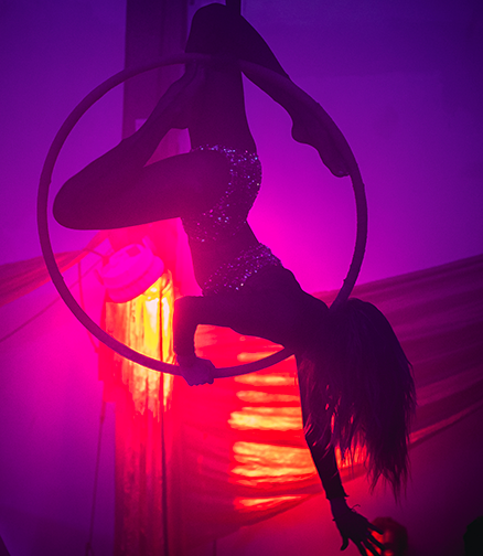 Silhouette d'une femme dans un cerceau aérien la tête en bas, sur fond lumineux chaud, teintes de rouge et mauve, durant une performance